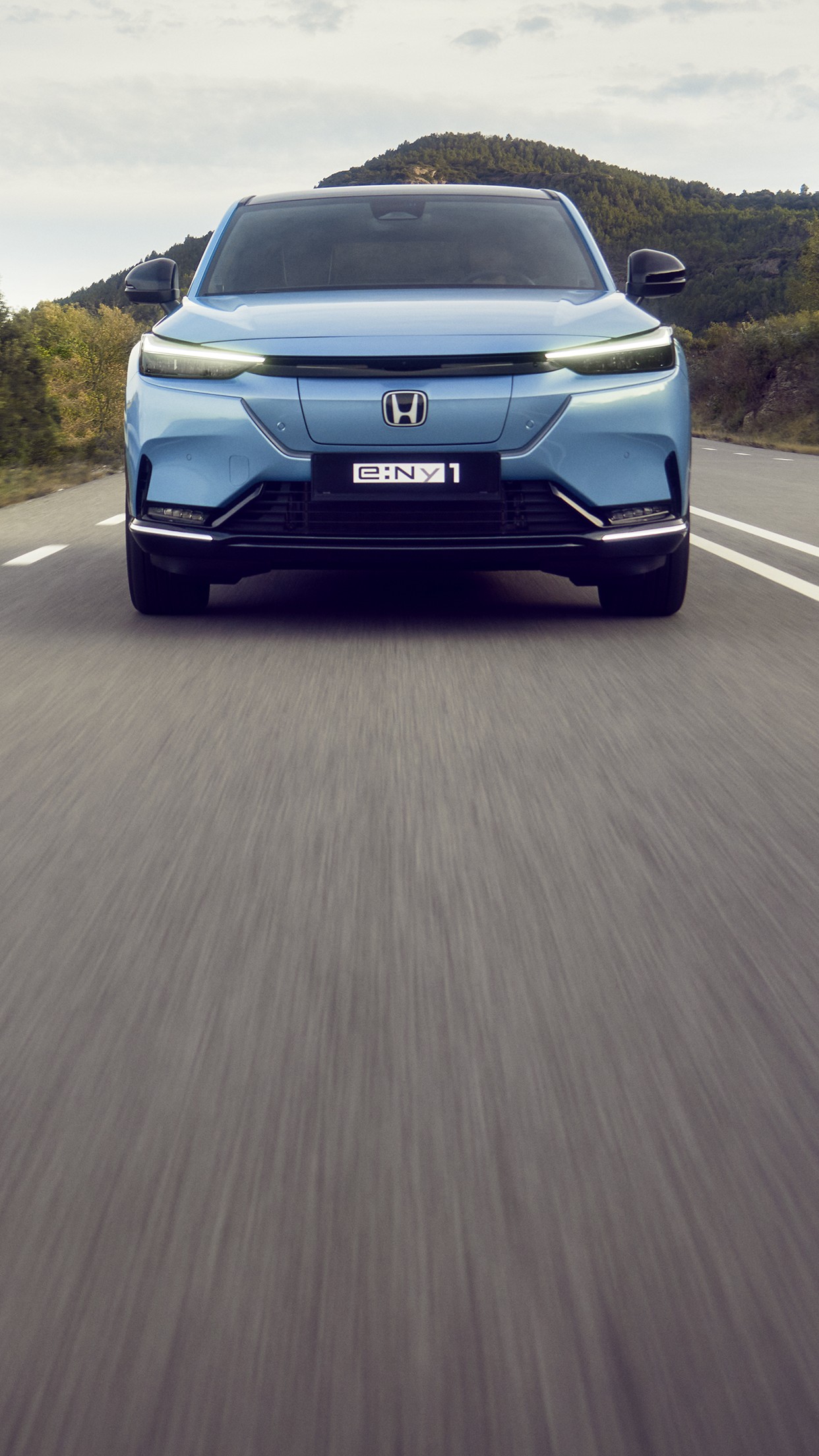 Honda e:Ny1 i Aqua Topaz Metallic kjører på en svingete vei med grønne åser i bakgrunnen.