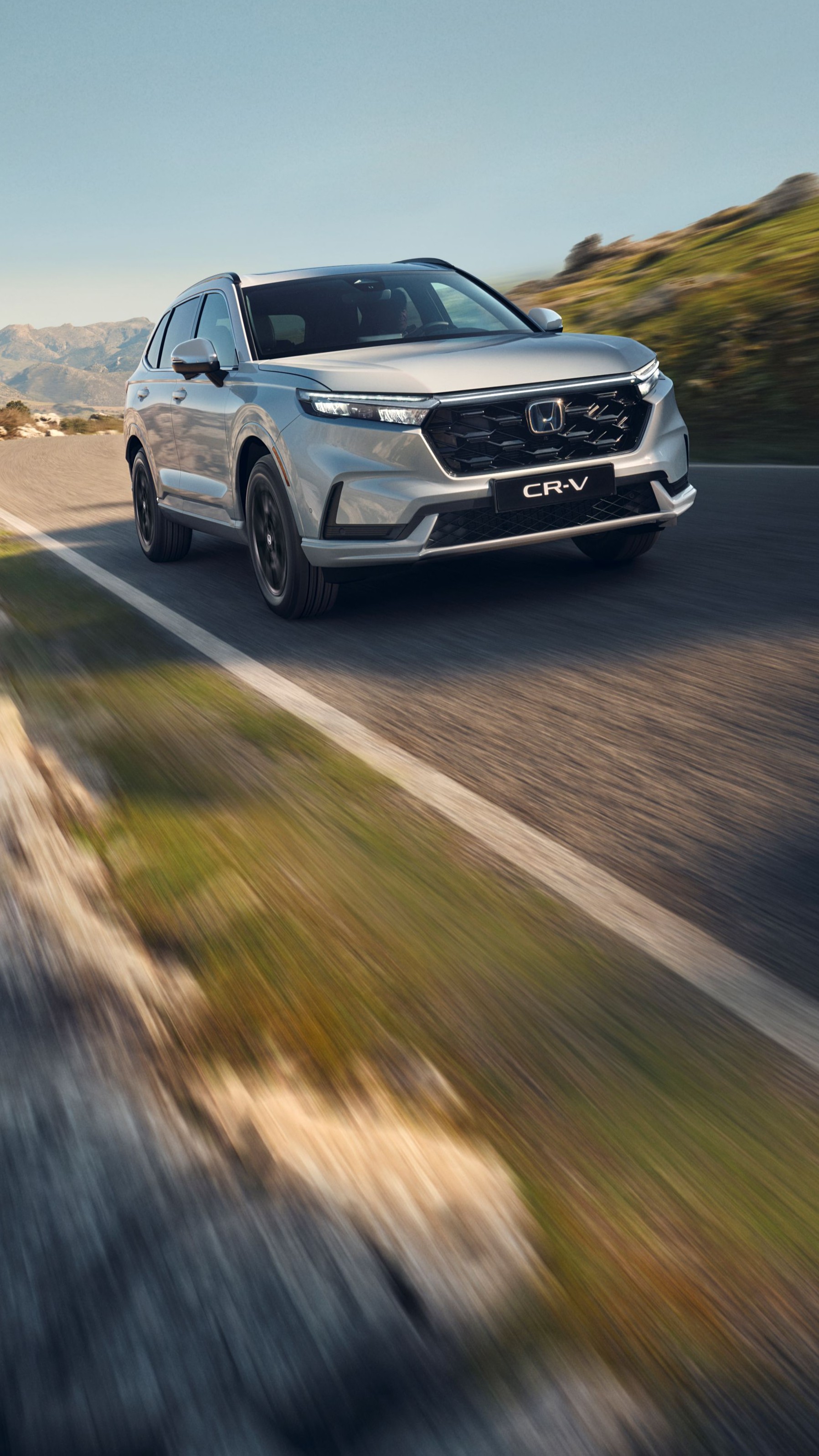 En sølvfarget Honda CR-V SUV kjører på en svingete fjellvei med steinete terreng og fjell i bakgrunnen. Kjøretøyet er i bevegelse, noe som skaper en følelse av fart og eventyr i det ville landskapet.