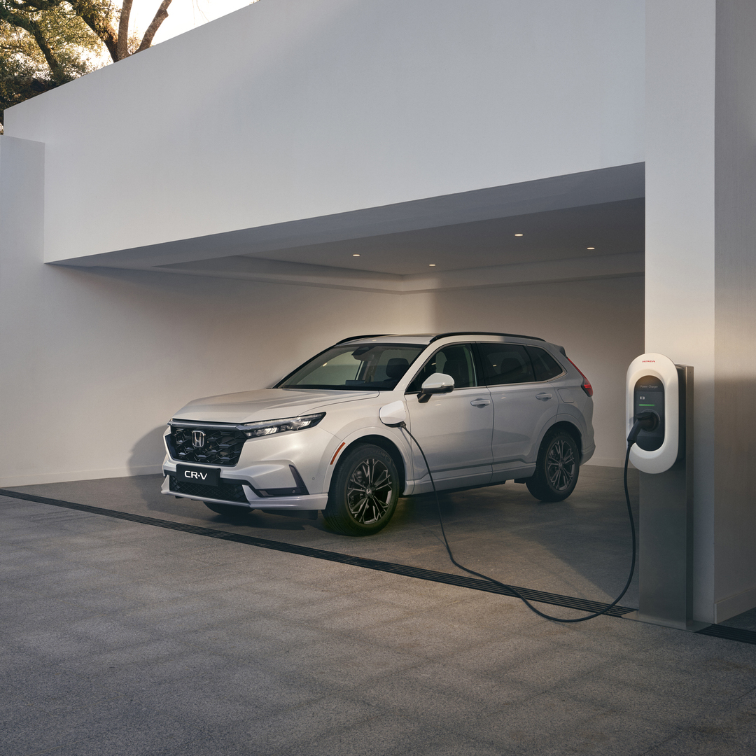 En hvit Honda CR-V plug-in hybrid står parkert i en minimalistisk hvit carport, koblet til en veggmontert elbillader.