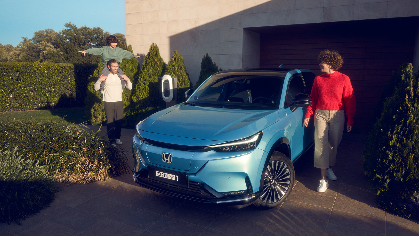 Familie som står ved siden av Honda e:Ny1 sett forfra parkert utenfor huset.