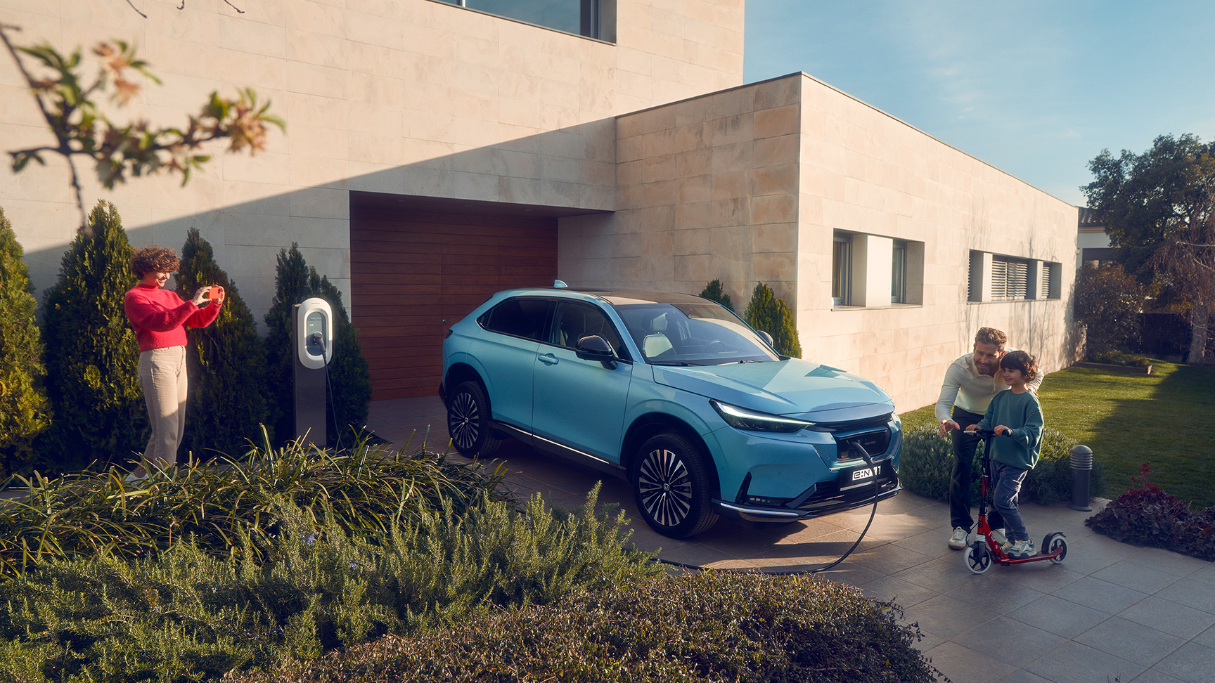 Familie som leker i oppkjørselen ved siden av Honda e:Ny1 som lader.