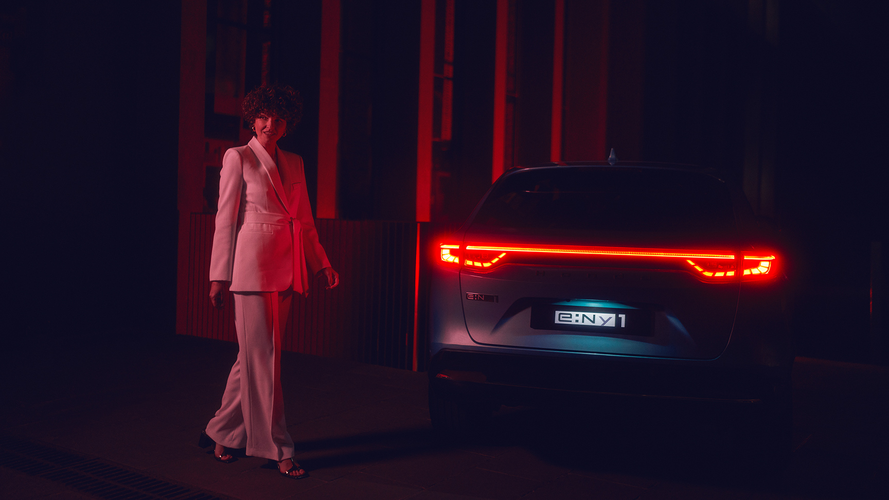 Modell som står ved siden av den opplyste baksiden av Honda e:Ny1 om kvelden.