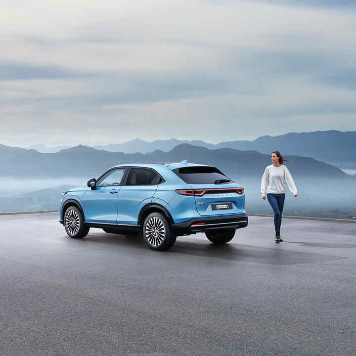 En blå Honda e:Ny1 parkert utendørs med utsikt over fjellene. En kvinne går ved siden av bilen.