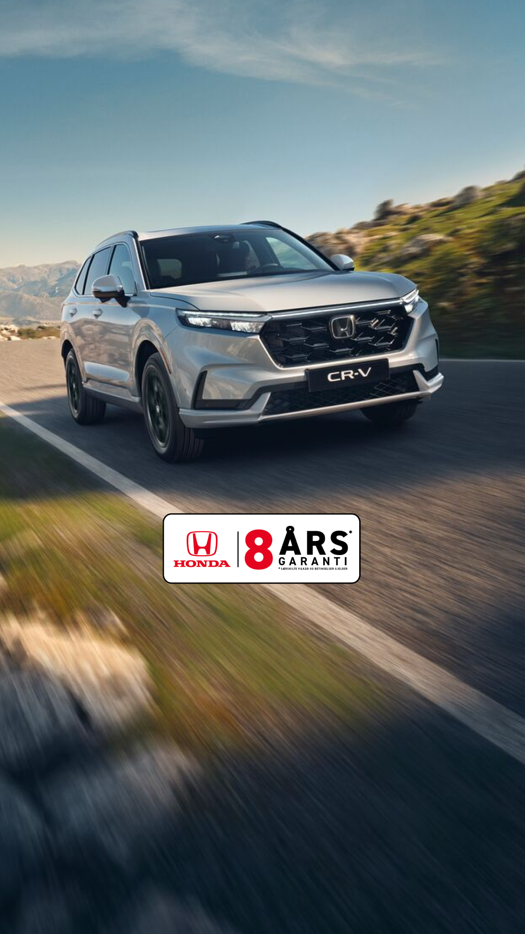 En sølvfarget Honda CR-V SUV kjører på en svingete fjellvei med steinete terreng og fjell i bakgrunnen. Kjøretøyet er i bevegelse, noe som skaper en følelse av fart og eventyr i det ville landskapet.