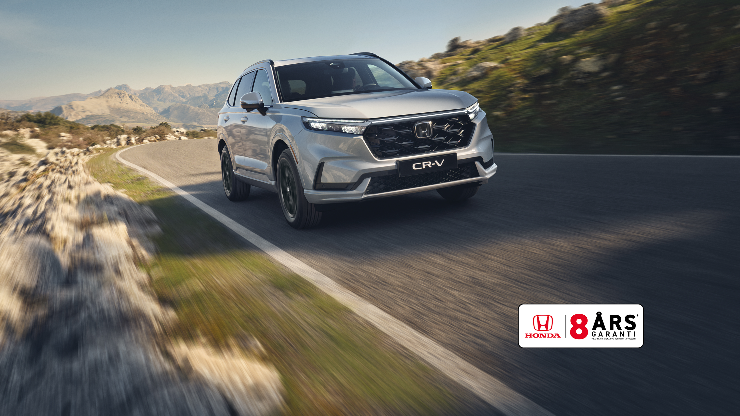En sølvfarget Honda CR-V SUV kjører på en svingete fjellvei med steinete terreng og fjell i bakgrunnen. Kjøretøyet er i bevegelse, noe som skaper en følelse av fart og eventyr i det ville landskapet.