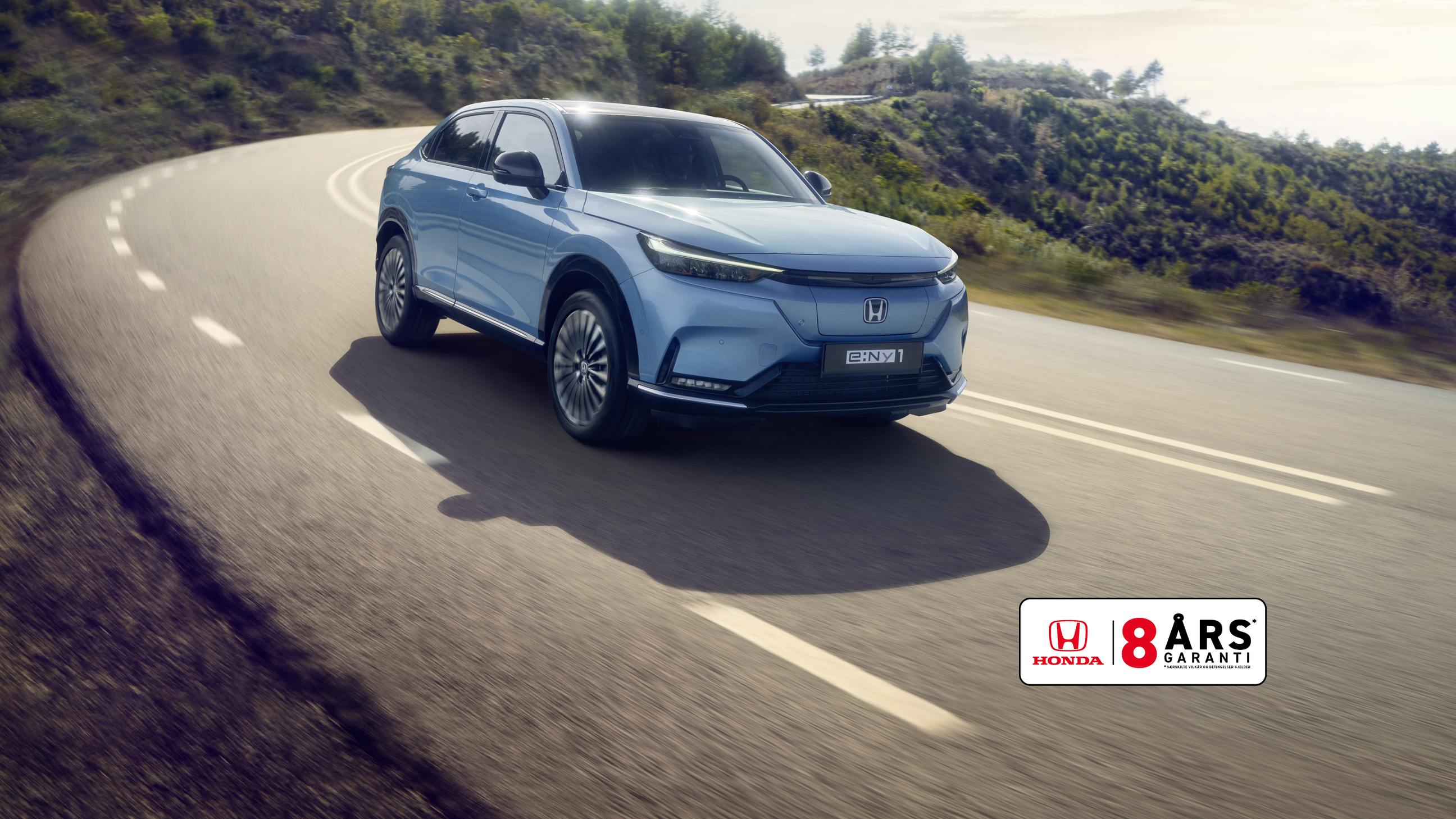 Honda e:Ny1 i Aqua Topaz Metallic kjører på en svingete vei med grønne åser i bakgrunnen.